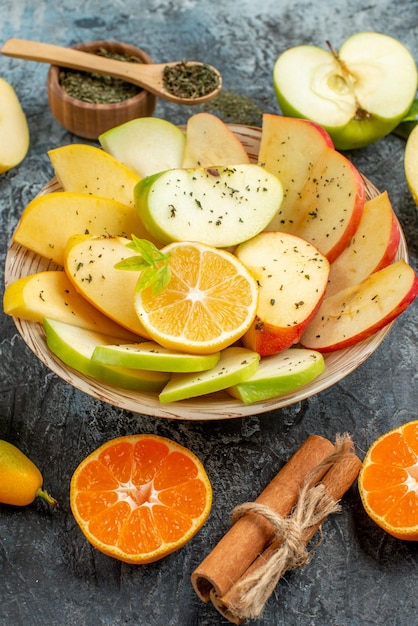 Вертикальный вид свежих натуральных кусочков яблока на белой тарелке с лимоном и корицей, лаймами, кумкватами, апельсинами на сером столе