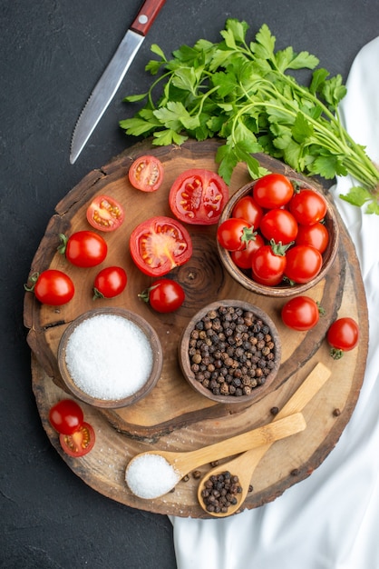 Vertical view of fresh tomatoes and spices in bowls spoons on wooden board on black surface