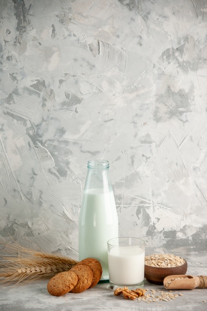 Vertical view of glass bottle and cup filled with milk on wooden tray and cookies spoon oats in brown pot on white table on ice background