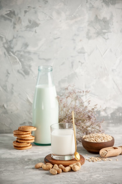 Vertical view of glass bottle and cup filled with milk on wooden tray and dry fruits stacked cookies spoon oats in brown pot on the left side on white table on ice background