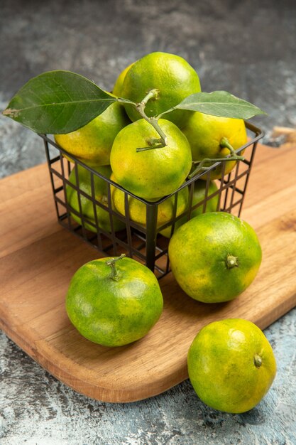 Vertical view of green mandarins with leaves inside and outside of a basket on wooden cutting board on gray table