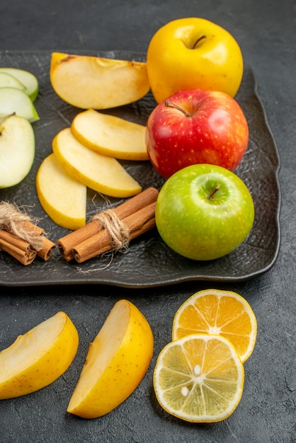 Vertical view of green yellow and red sliced and whole fresh apples on a black plate and cinnamon limes on a dark table