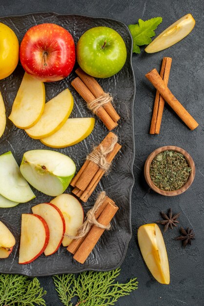 Vertical view of green yellow and red sliced and whole fresh apples on a black tray and cinnamon limes on a dark table