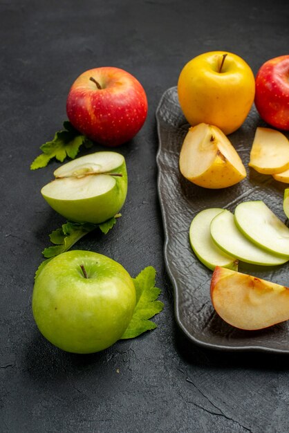 Vertical view of green yellow and red sliced and whole fresh apples on a black tray and cinnamon limes on a dark table