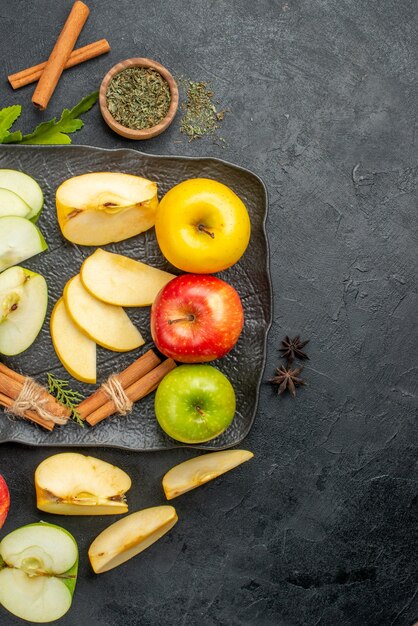 Vertical view of green yellow and red sliced and whole fresh apples on a black tray and cinnamon limes on the right side on a dark background