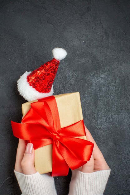 Vertical view of hand holding beautiful gift with bow-shaped ribbon next to santa claus hat on dark background