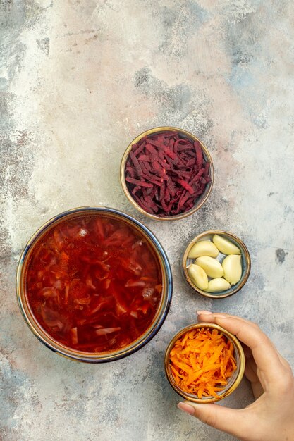 Vertical view of hand holding chopped carrots in a white small plate a blue pot with delicious borscht soup and other necessary vegetables on a colorful background