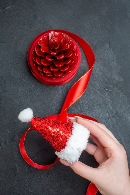 Vertical view of hand holding santa claus hat and red conifer cone on dark background