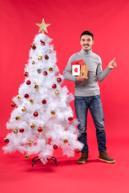 Вертикальный вид красивого человека, стоящего возле украшенной белой рождественской елки и держащего подарки