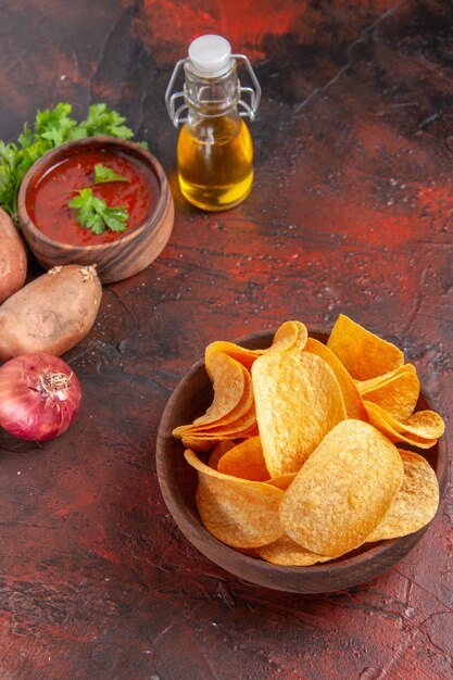 Vertical view of homemade delicious crispy chips in a small brown bowl garlic green ketchup potatoes onion oil bottle on dark table