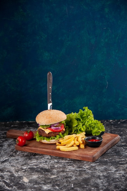 Vertical view of knife in meat sandwich and fries tomatoes with stem pepper on wooden board ketchup on dark blue surface