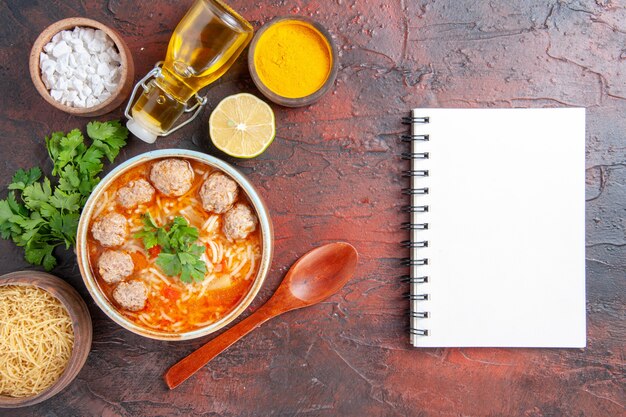 Vertical view of meatballs soup with noodles in a brown bowl lemon spoon a bunch of green and oil bottle pasta and notebook on dark table