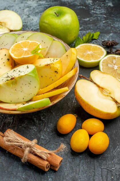 Вертикальный вид натуральных яблок с лимоном на белой тарелке и корицей, лаймов, кумкватов, апельсинов на темном столе