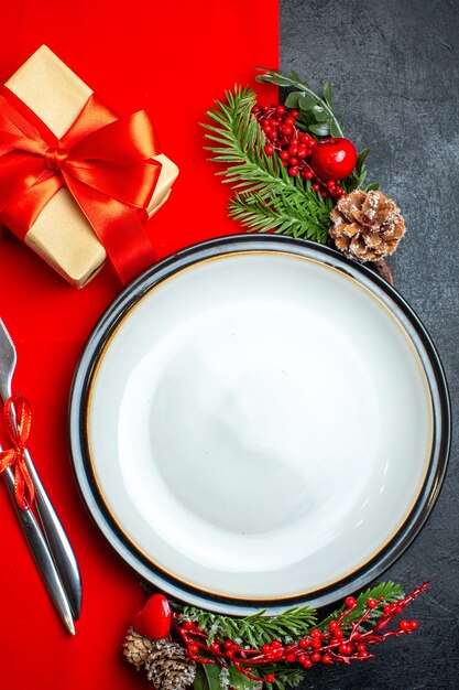 Вертикальный вид новогоднего фона с обеденной тарелкой, набор столовых приборов, украшения, аксессуары, еловые ветки, рядом с подарком на красной салфетке