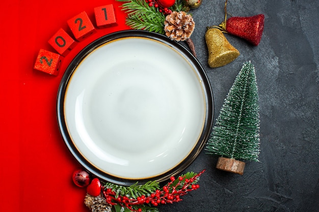 Vista verticale dello sfondo del nuovo anno con la decorazione della piastra della cena accessori rami di abete e numeri su un tovagliolo rosso accanto all'albero di natale su una tavola nera