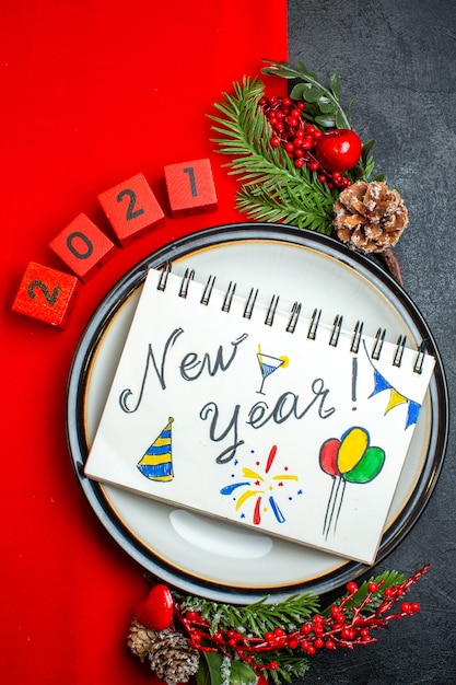 Vertical view of New year background with notebook with new year drawings on a dinner plate decoration accessories fir branches and numbers on a red napkin on a black table