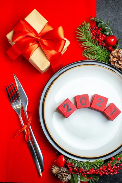 Vertical view of New year background with numbers on dinner plate cutlery set decoration accessories fir branches next to a gift on a red napkin