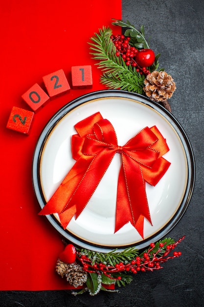 Вертикальный вид новогоднего фона с красной лентой на аксессуарах для украшения обеденной тарелки еловые ветки и числа на красной салфетке на черном столе