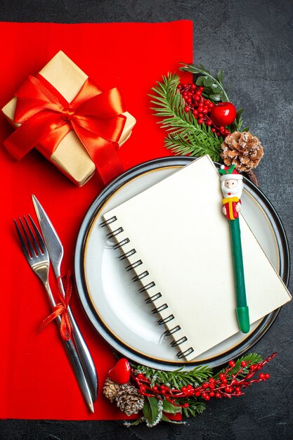 Вертикальный вид новогоднего фона со спиральной записной книжкой на обеденной тарелке, набор столовых приборов, украшения, аксессуары, еловые ветки, рядом с подарком на красной салфетке