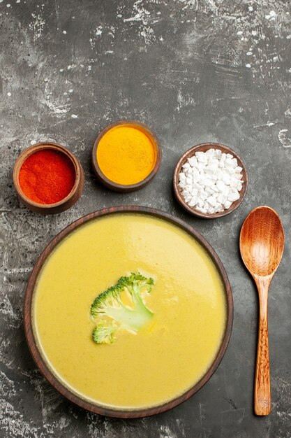 Бесплатное фото Вертикальный вид сливочного супа из брокколи в коричневой миске с различными специями и ложкой на сером столе
