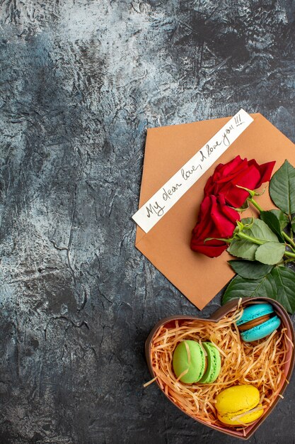 Vertical view of red roses and envelope with love letter and different macarons on icy dark background