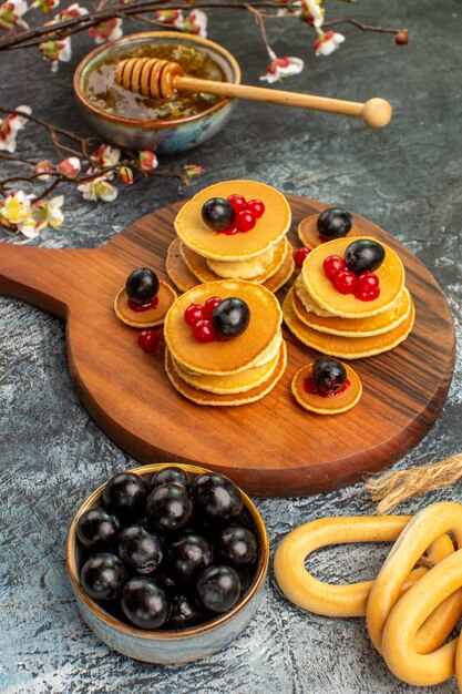 Вертикальный вид кольцевого печенья, фруктовых блинов, меда в миске и черной вишни на сером столе