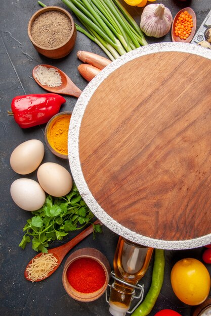 Vertical view of round cutting board among fresh vegetables lemon fallen oil bottle eggs different spices on black