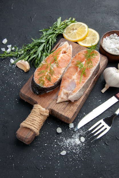 Vertical view salmon fish on brown wooden cutting board with green and cutlery set garlic salt on dark background