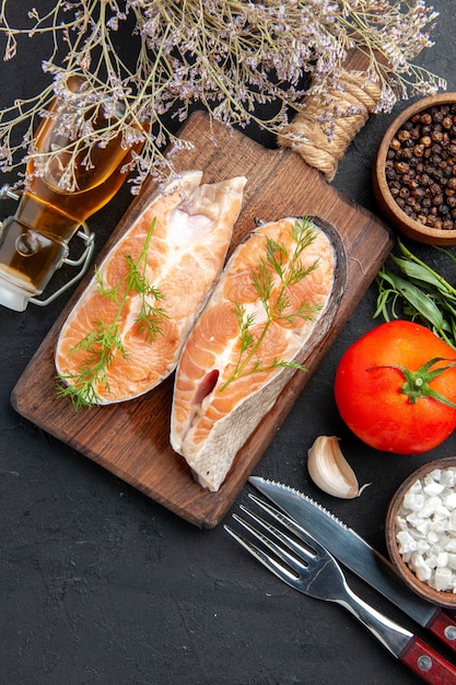 Vertical view of salmon fish on brown wooden cutting board with green pepper fir branches and cutlery set fallen oil bottle tomato salt pepper on dark table
