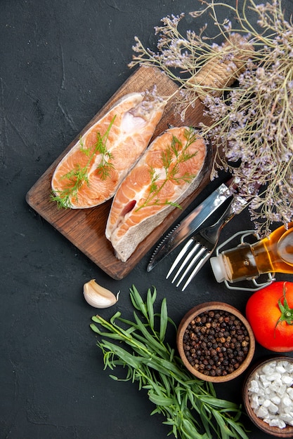 Vertical view of salmon fish on brown wooden cutting board with green pepper fir branches and cutlery set fallen oil bottle tomato salt pepper on dark table