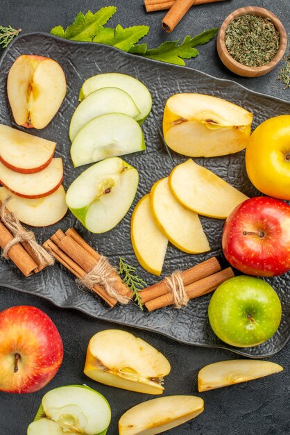 Vertical view of several types of sliced and whole fresh apples on a black tray and cinnamon limes on a dark background