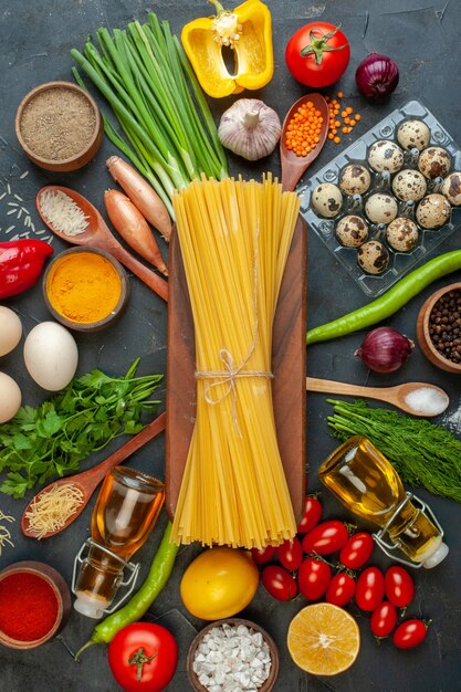 Vertical view of spagetti on a wooden board among organic vegetables eggs fallen oil bottle pepper on black
