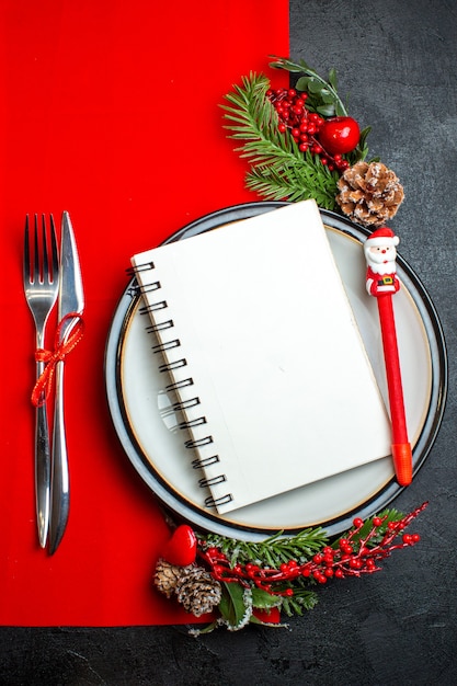 Vertical view of a spiral notebook and a pen on dinner plate with decoration accessories fir branches and cutlery set on a red napkin