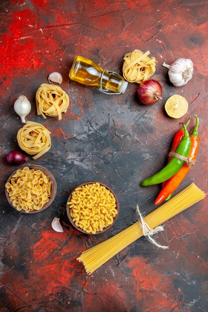 Vista verticale della gustosa preparazione per la cena con pasta cruda in varie forme e aglio olio caduto bottiglia aglio limone su sfondo di colore misto