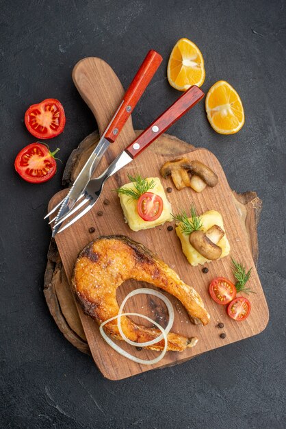 Vertical view of tasty fried fish and mushrooms tomatoes greens on cutting board cutlery set pepper on black surface