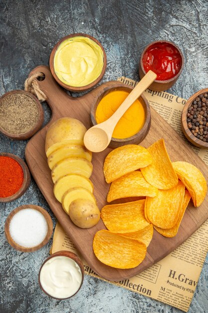 Vertical view of tasty homemade chips cut potato slices on wooden cutting board and different spices on newspaper on gray background