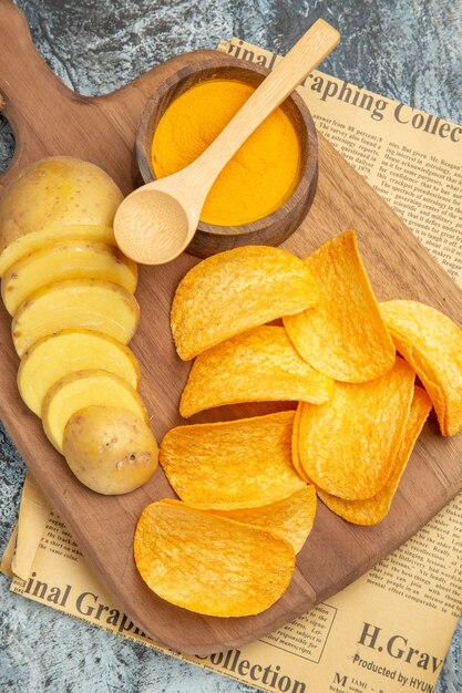 Vertical view of tasty homemade chips cut potato slices on wooden cutting board on newspaper on gray background