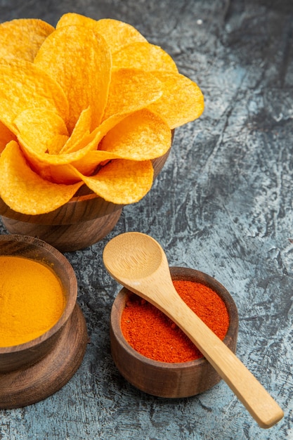 Vertical view of tasty potato chips decorated like flower shaped different spices with spoons on them on gray table