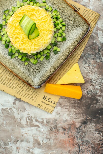 Vertical view of tasty salad served with chopped cucumber on an old newspaper and two kinds of cheese on mixed color background