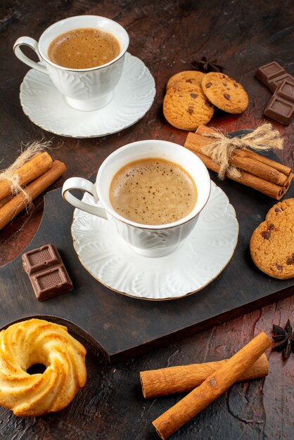 Vertical view of two cups of coffee cookies cinnamon limes chocolate bars on wooden cutting board on dark background