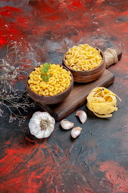 Vertical view of various types of uncooked pastas on wooden cutting board and garlic on mixed color background