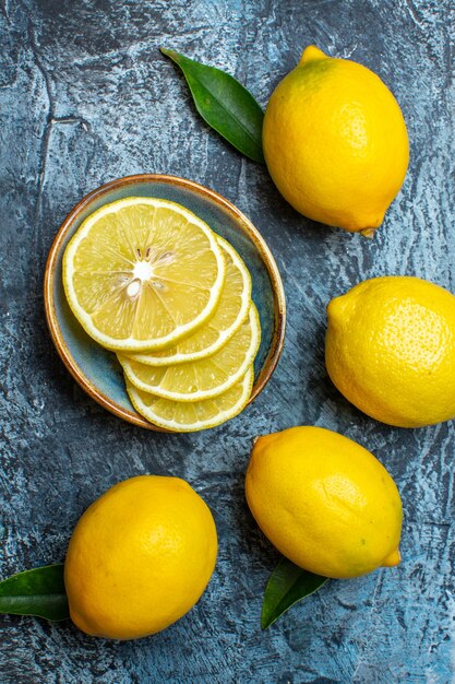 Вертикальный вид целых и нарезанных свежих лимонов с листьями слева на темном фоне