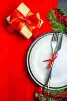 Foto gratuita vista verticale dello sfondo di natale con set di posate con nastro rosso su un piatto da pranzo accessori per la decorazione rami di abete accanto a un regalo su un tovagliolo rosso