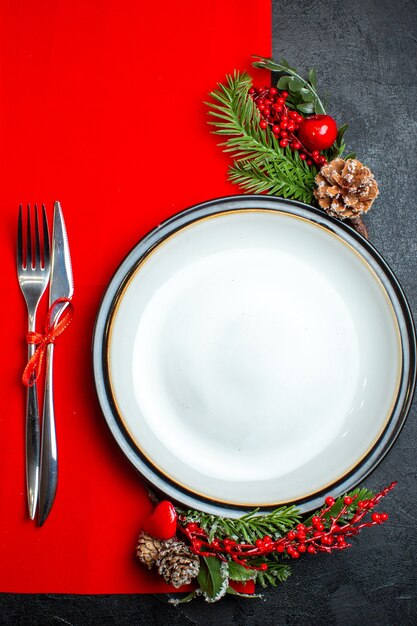 Вертикальный вид на рождественский фон с аксессуарами для украшения обеденной тарелки, еловыми ветками и набором столовых приборов на красной салфетке