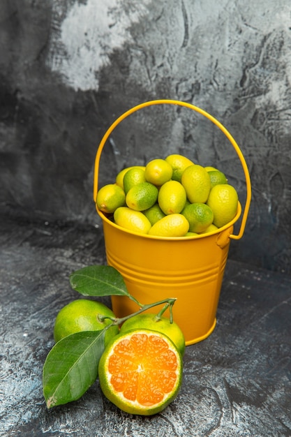 Vertical view of a yellow bucket full of fresh green tangerines and cut in half tangerines on gray background