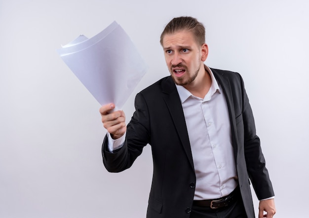 Very angry handsome business man wearing suit holding blank pages gesturing with aggressive expression standing over white background