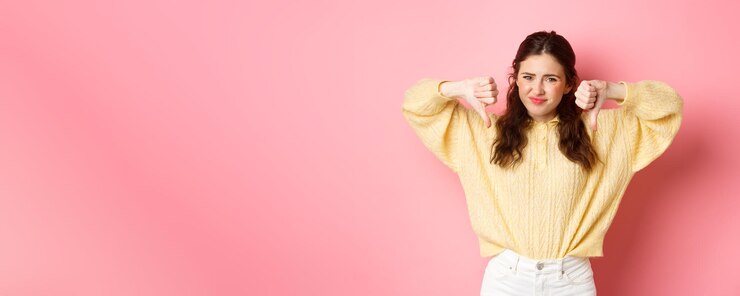 無料Photo 非常に悪い がっかりした若い女性が何かひどいことに不平を言い、親指を下にして嫌悪感を示し、顔をしかめ、顔をしかめ、頭を振って否定的なフィードバックを与える ピンクの背景