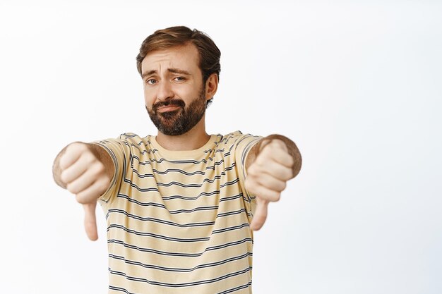 Very bad Portrait of bearded guy shows thumbs down and smirks with dislike disappointed by smth standing over white background
