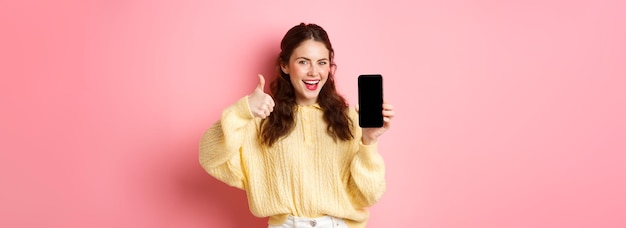 Free photo very good app smiling satisfied woman showing thumbs up and empty smartphone screen recommending app