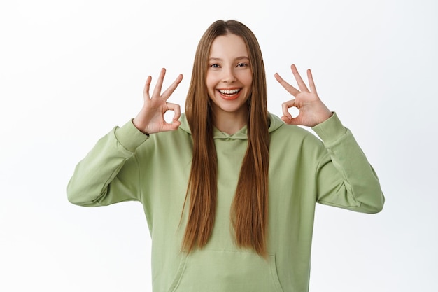 Molto bene. bella studentessa, adolescente mostra il segno giusto e dice di sì, sorridendo soddisfatto, approva e mi piace, loda la buona scelta, ben fatto, bel lavoro, in piedi su sfondo bianco.